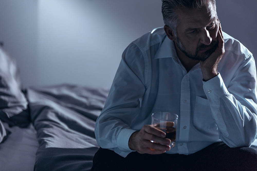 Close-up of a man in crisis worrying about his relationship - sitting on a bed with one hand on his chin and holding a glass of alcohol in the other - Ryan Family Law